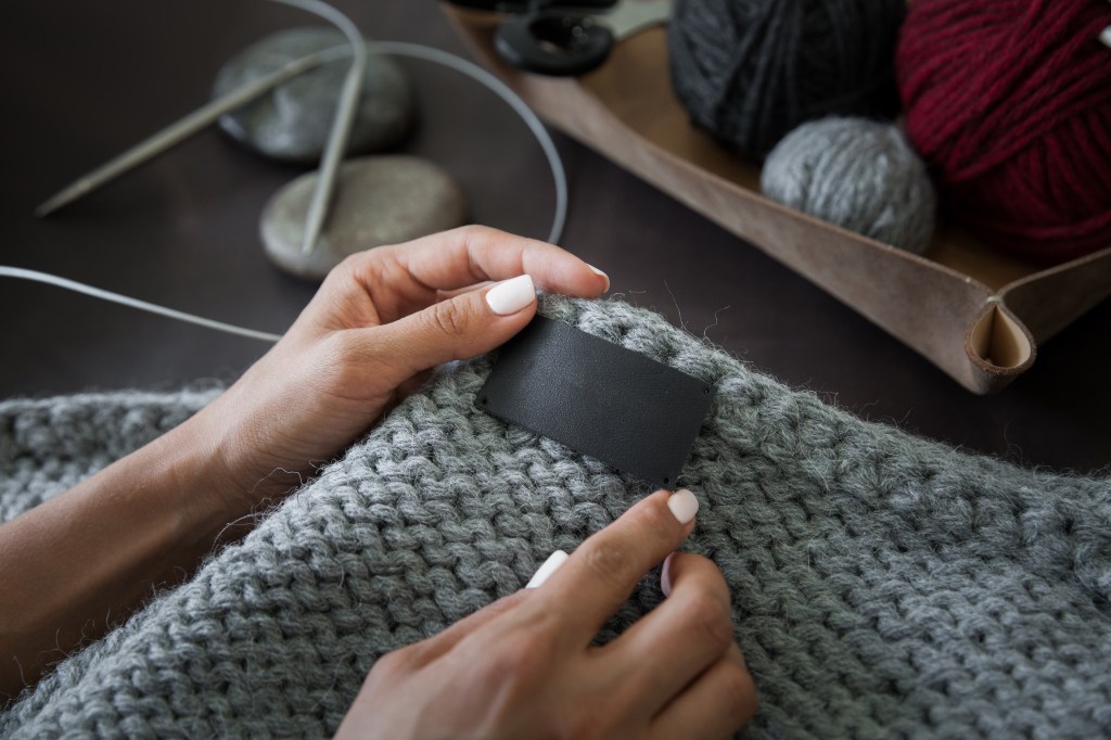 Woman removing blank clothing label on knitted clothing. Closeup, hands, sweater, balls of yarn and knitting needles in image.